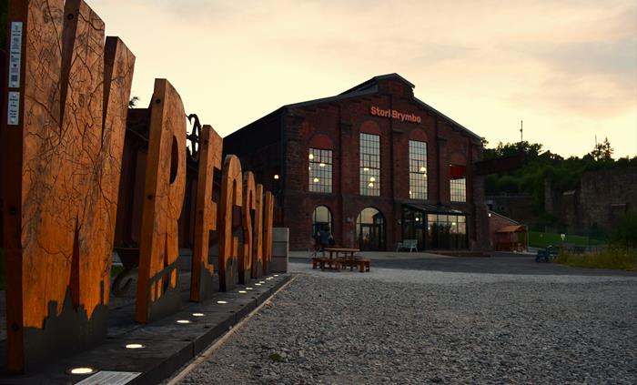 Wooden letters spelling Wrecsam in front of a renovated industrial machine shop-turned-coffeehouse at sunset
